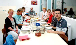 Foto von der Kochgruppe, die sich an einem großen Esstisch versammelt hat. Kommentar: 'Alle sitzen am gedeckten Tisch, gleich kann das gemeinsam zubereitete Mahl beginnen...'